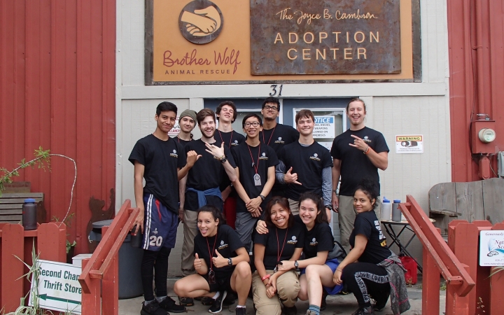 A group of students smile at the camera on the steps of a building under a sign. The sign reads "Brother Wolf Animal Rescue, Adoption Center." Additional text is not readable. 